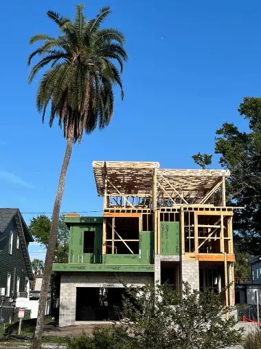 Modern home framed in green sheathing under a tall palm tree, upper floor deck still open